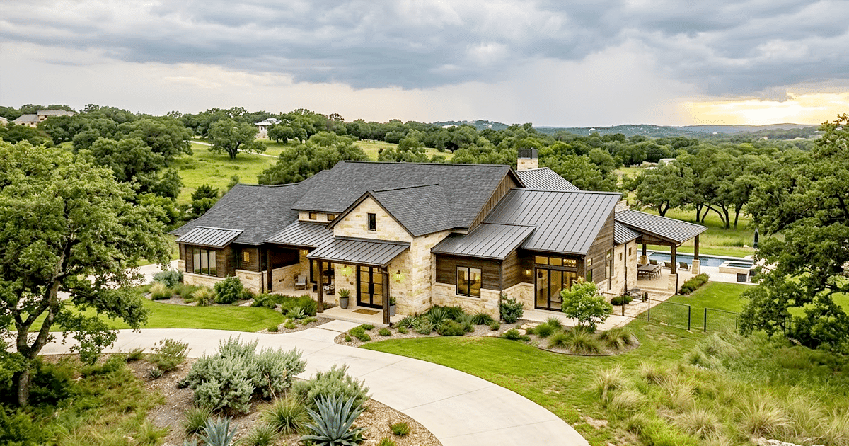 An aerial view of a large, luxurious ranch-style home in the Texas Hill Country near New Braunfels, demonstrating the use of durable residential roofing materials. The roof is a combination of dark, standing seam metal panels and asphalt shingles, expertly installed by professional roofers. The house is surrounded by a sprawling green lawn, native oak trees, and curated landscaping with agave plants under a dramatic, cloudy sky, highlighting the craftsmanship of New Braunfels roofers.