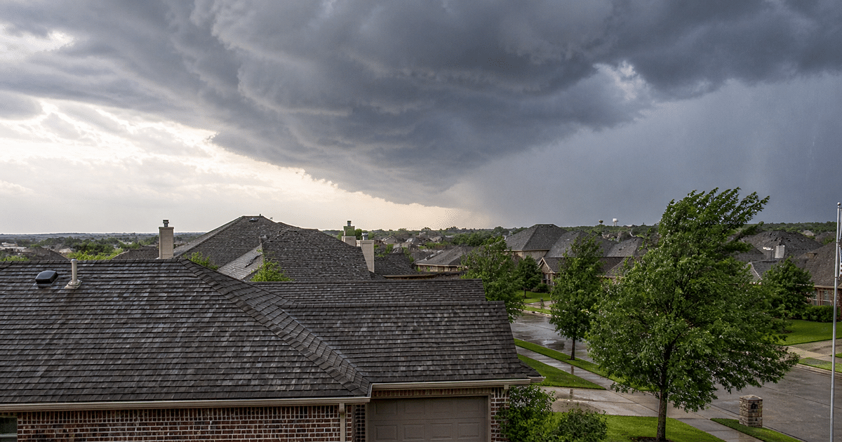 Dallas Fort Worth residential neighborhood as a hail storm approaches, showing shingle rooftops, dark storm clouds, and light rain moving into the area
